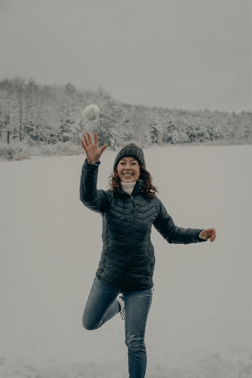 Smilende kvinne  med lue, jakke og jeans kaster snøball mot fotografen