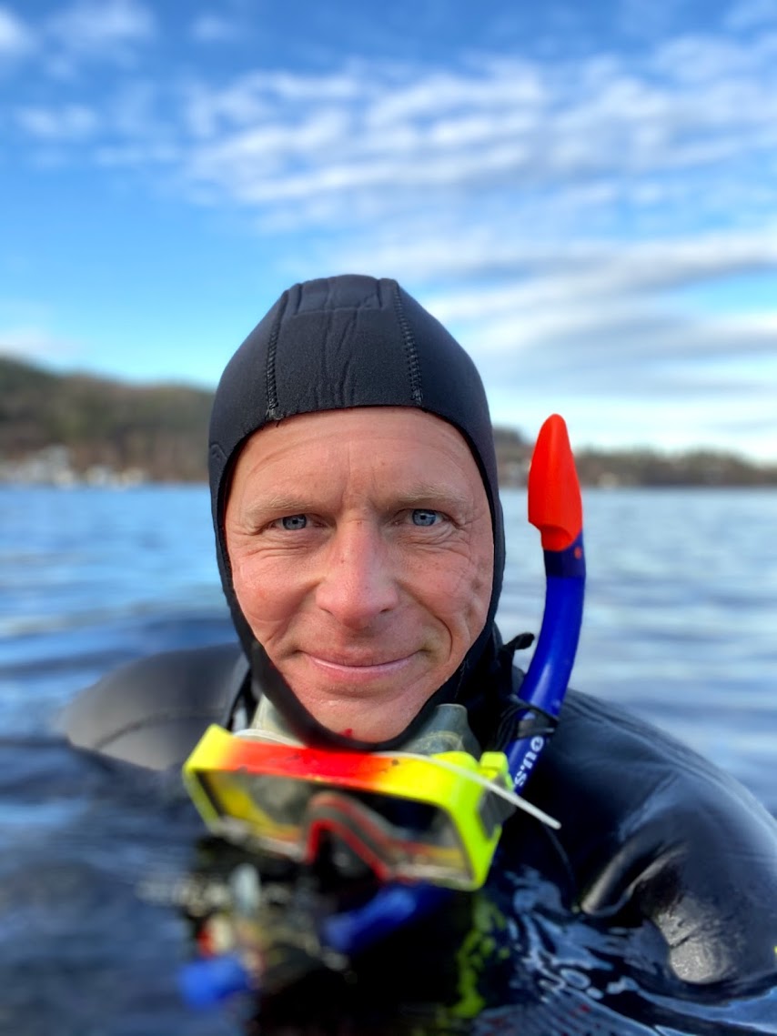 Smilende mann i våtdrakt med gule dykkerbriller stikker hodet opp av vannskorpa